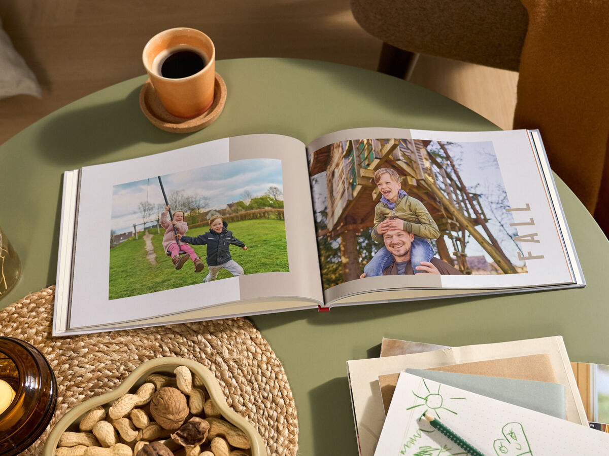 Aufgeschlagenes CEWE FOTOBUCH mit herbstlichen Familienfotos und der Aufschrift „FALL“ auf einem grünen Tisch; daneben stehen eine Tasse Kaf