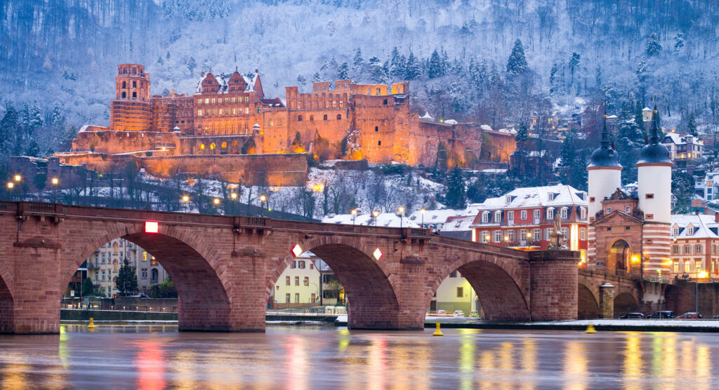 Heidelberger Schloss im Winter. Im Vordergrund die Karl-Theodor-Brücke und der Neckar, in dem sich die Lichter der Stadt spiegeln.