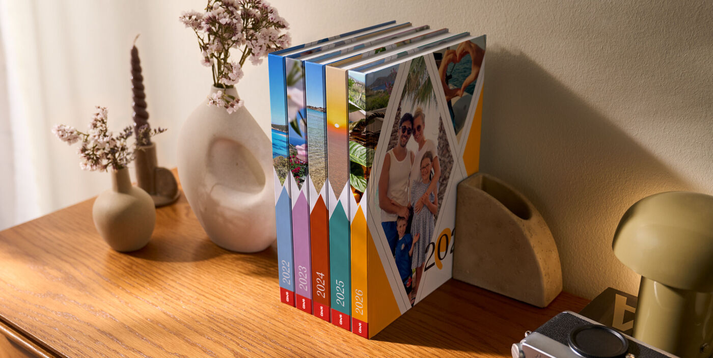 Mehrere bunte Bücher im einheitlichen Einband stehen aufrecht auf einem Holzregal neben dekorativen Vasen