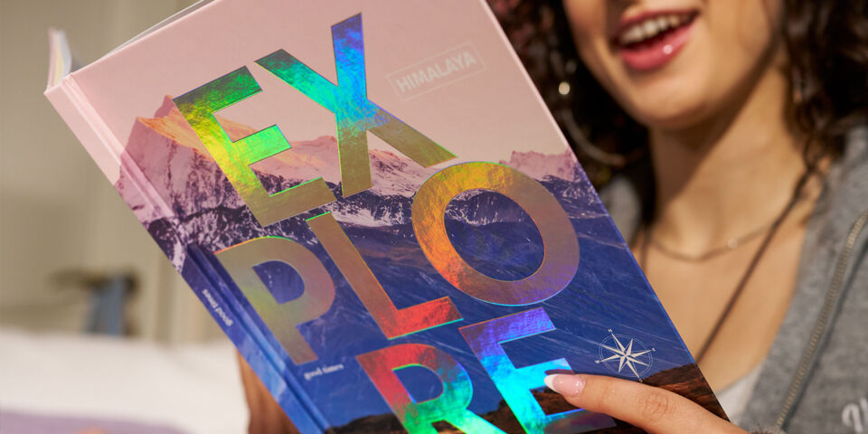 Lachende Person hält ein CEWE FOTOBUCH mit Bergmotiv und dem Schriftzug „Explore“ mit Veredelung mit holografischem Effekt