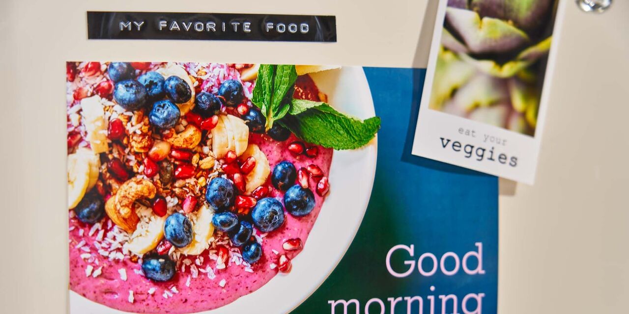 An einem cremefarbenen Kühlschrank hängen zwei Sofortfotos in unterschiedlichen Formaten. Das größere zeigt eine Frühstücks-Bowl und die Aufschrift „Good morning“. Ein kleineres Foto zeigt die Nahaufnahme einer Artischocke sowie die Aufschrift „Eat your veggies“. Über den Fotos befindet sich ein geprägtes Etikett mit dem Text „My favorite food“.