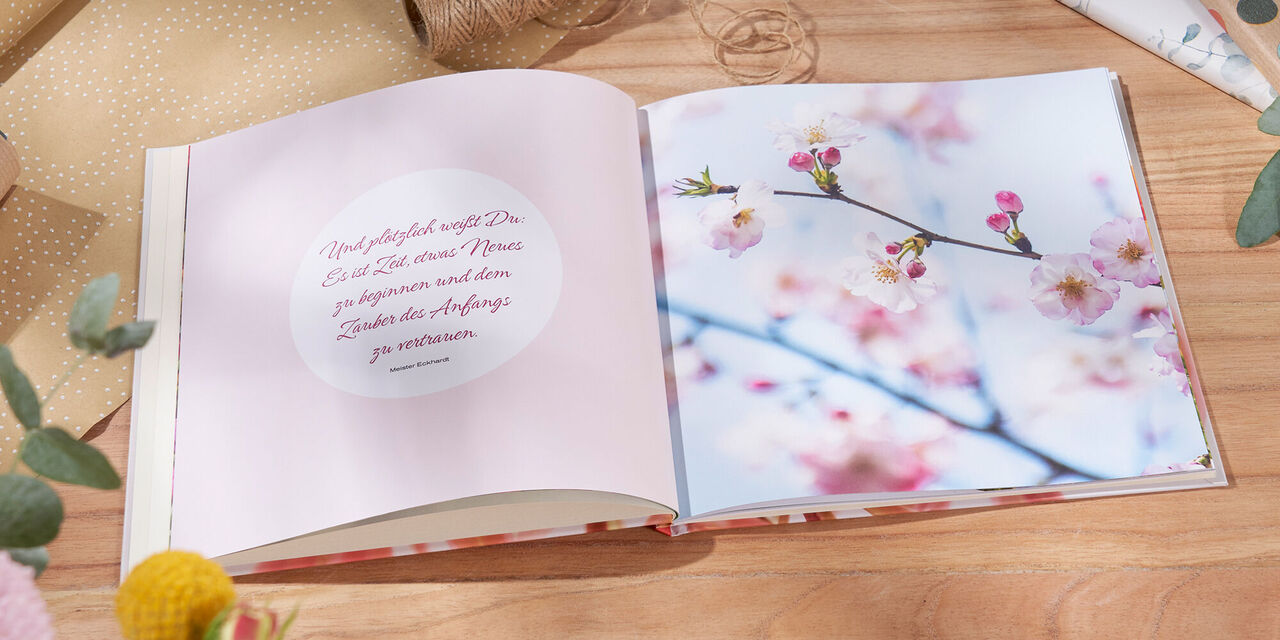 Auf einem Tisch liegt ein aufgeschlagenes CEWE FOTOBUCH. Daneben liegen Geschenkpapier und Blumen. Auf der Doppelseite sind eine Nahaufnahme von Apfelblüten und ein rundes Clipart mit einem Spruch zu sehen.