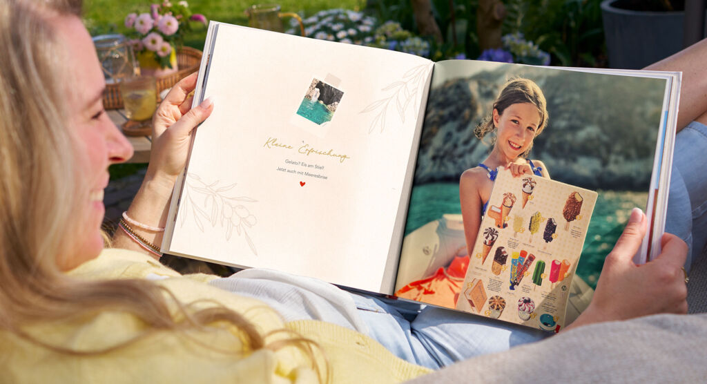 Eine Frau sitzt entspannt im Garten und sieht sich ein Fotobuch an, das sommerliche Urlaubserinnerungen zeigt.