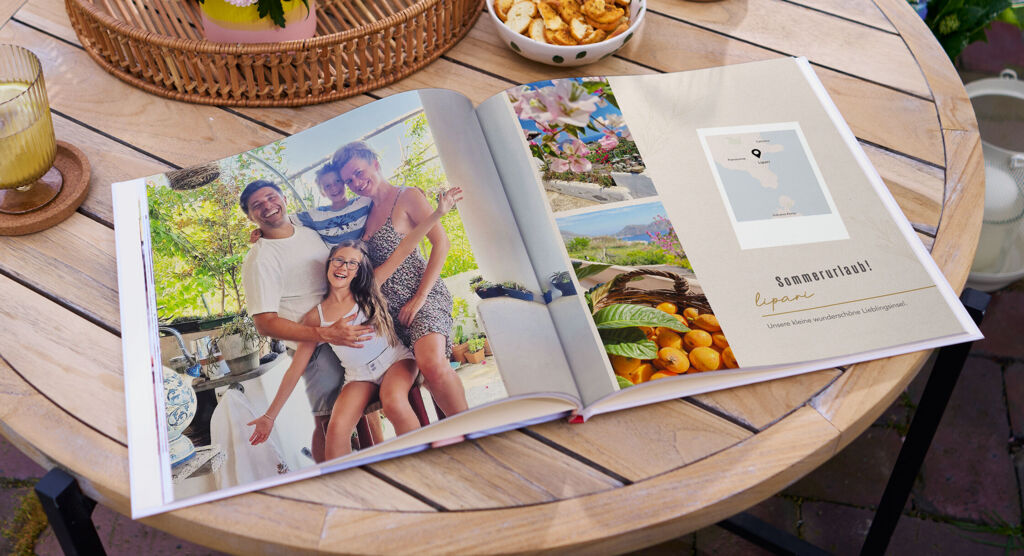 Ein runder Holztisch im Garten mit einem geöffneten Fotobuch, Getränken, Snacks und einem Blumenstrauß auf einem Tablett.