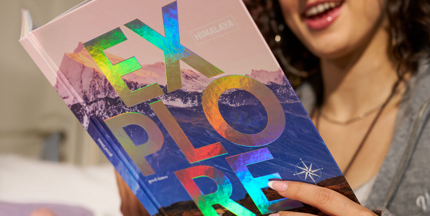 Lachende Person hält ein CEWE FOTOBUCH mit Bergmotiv und dem Schriftzug „Explore“ mit Veredelung mit holografischem Effekt