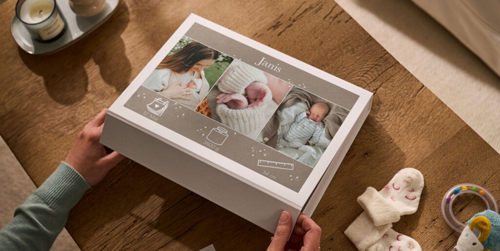 Person öffnet eine weiße Erinnerungsbox mit drei Babyfotos, dem Namen „Janis“ und kleinen Erinnerungen auf einem Holztisch.